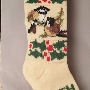 May include: A white knitted Christmas stocking with a red cuff, featuring a green holly design and two brown and white birds. The stocking is personalized with the name "BETH" in green yarn. The label reads "Bernat Knitted Stocking W00630 Chickadees".