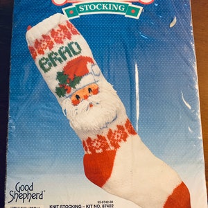 May include: Knitting kit for a Christmas stocking featuring a Santa Claus design. The kit includes yarn, needles, and instructions. The stocking is white with red accents and a green holly sprig. The kit is labeled "Happy Santa" and "Knit Stocking - Kit No. 87402".