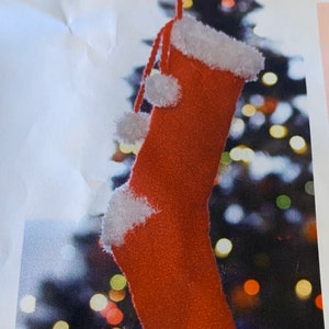 May include: A red and white knitted Christmas stocking with a pom-pom on top. The stocking is hanging from a string and is in front of a blurry background of Christmas lights. The label reads "No. 98862P BERNAT® CRAFT FAIR CHRISTMAS STOCKING | Kn"