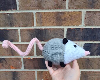 Könnte beinhalten: Ein gestrickter grauer und weißer Opossum mit einem rosa Schwanz. Der Opossum hat schwarze Augen und eine schwarze Nase.
