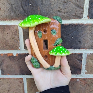 Puede incluir: Una cubierta de interruptor de luz de cerámica marrón con dos acentos de hongos verdes y blancos. La cubierta tiene una abertura rectangular para el interruptor y dos aberturas circulares. Las setas tienen lunares blancos.