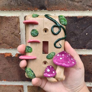 Op de afbeelding: Een decoratieve lichtschakelaar met een grillig paddenstoelontwerp. De afdekking is bruin met roze en groene accenten, waaronder paddenstoelkapjes met witte stippen en getextureerd groen mos. Een donkergroen, wijnstokachtig element voegt details toe.