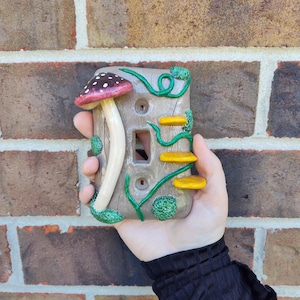 Op de afbeelding: Een decoratieve lichtschakelaar cover met een grillig paddestoelontwerp. De cover is geschilderd in bruin, rood, wit, geel en groen. De paddestoel heeft een rode hoed met witte stippen. De cover wordt in een hand gehouden.