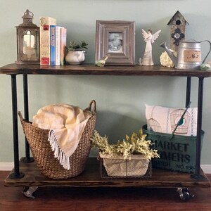May include: A rustic industrial style console table with a dark wood top and black metal legs. The table has two shelves and is decorated with a variety of items, including books, a lantern, a picture frame, a watering can, and a birdhouse. The table is on wheels and is perfect for a hallway or entryway.