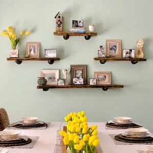 Puede incluir: Un conjunto de tres estantes de madera con un acabado r&uacute;stico, decorados con fotos enmarcadas, una casita de p&aacute;jaros y un jarr&oacute;n de tulipanes amarillos. Los estantes est&aacute;n montados en una pared verde claro. La palabra "LOVE" es visible en un peque&ntilde;o letrero de madera.