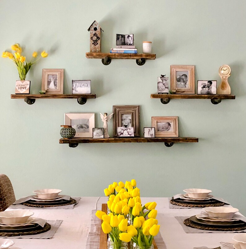 Puede incluir: Un conjunto de tres estantes de madera con un acabado r&uacute;stico, decorados con fotos enmarcadas, una casita de p&aacute;jaros y un jarr&oacute;n de tulipanes amarillos. Los estantes est&aacute;n montados en una pared verde claro. La palabra "LOVE" es visible en un peque&ntilde;o letrero de madera.
