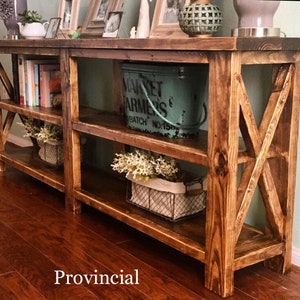 May include: A rustic wooden console table with two shelves and an X-shaped design on the legs. The table is stained a dark brown color and has a distressed finish. The table is decorated with a vintage metal bucket, a small plant, and a book.