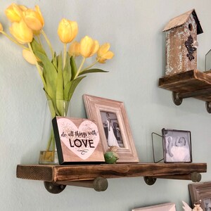 Puede incluir: Una estanter&iacute;a de madera r&uacute;stica con soportes met&aacute;licos, que presenta una foto enmarcada, un peque&ntilde;o letrero de madera con el texto "do all things with LOVE", una casita de p&aacute;jaros y un jarr&oacute;n de tulipanes amarillos.