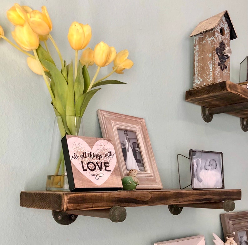 Puede incluir: Una estanter&iacute;a de madera r&uacute;stica con soportes met&aacute;licos, que presenta una foto enmarcada, un peque&ntilde;o letrero de madera con el texto "do all things with LOVE", una casita de p&aacute;jaros y un jarr&oacute;n de tulipanes amarillos.