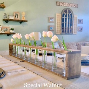 May include: A wooden centerpiece with a rustic finish, featuring a row of glass tubes for holding flowers. The centerpiece is labeled "Special Walnut" and is displayed on a white tablecloth with pink tulips in the tubes.