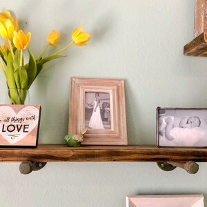 Puede incluir: Una estanter&iacute;a de madera r&uacute;stica con dos soportes met&aacute;licos, que sostiene una foto en blanco y negro enmarcada, un peque&ntilde;o letrero de madera con el texto "do all things with LOVE" y una figurita de p&aacute;jaro verde. La estanter&iacute;a est&aacute; montada en una pared verde claro.