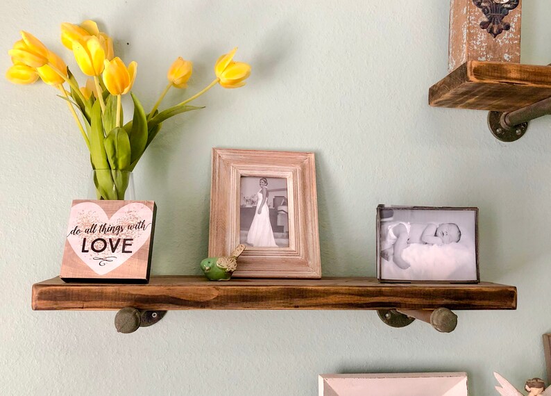 Puede incluir: Una estanter&iacute;a de madera r&uacute;stica con dos soportes met&aacute;licos, que sostiene una foto en blanco y negro enmarcada, un peque&ntilde;o letrero de madera con el texto "do all things with LOVE" y una figurita de p&aacute;jaro verde. La estanter&iacute;a est&aacute; montada en una pared verde claro.