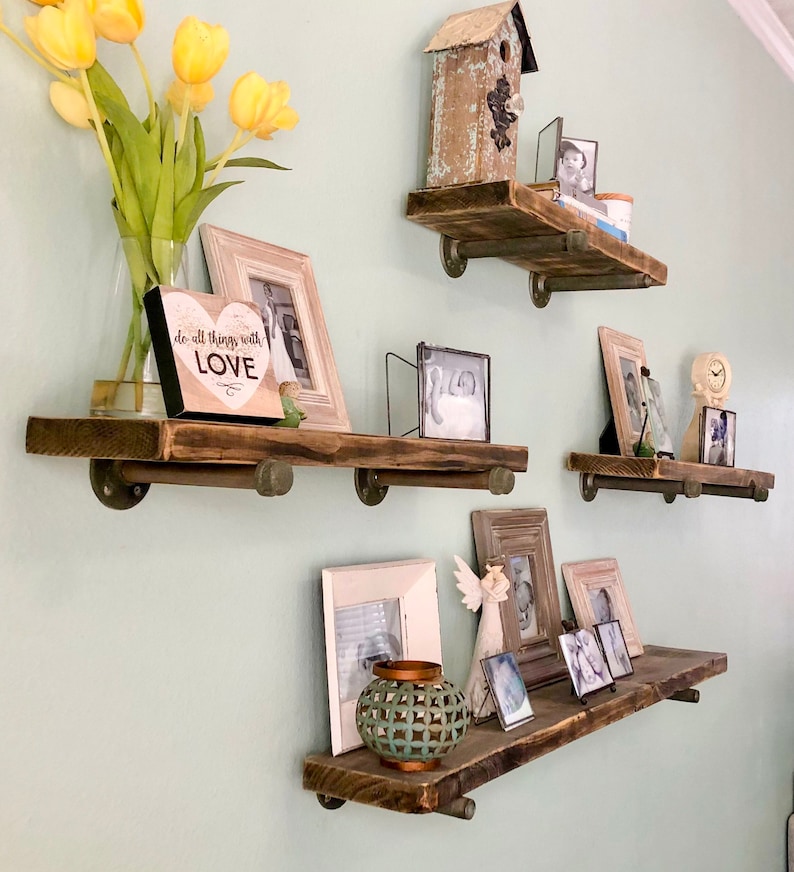Puede incluir: Cuatro estantes de madera r&uacute;stica con soportes de tuber&iacute;a met&aacute;lica, decorados con fotos enmarcadas, una peque&ntilde;a casita de p&aacute;jaros, una placa decorativa en forma de coraz&oacute;n que dice "do all things with love" y una peque&ntilde;a figurita de &aacute;ngel decorativa.