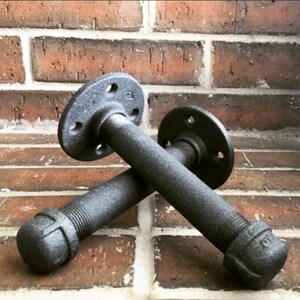 Puede incluir: Dos soportes de estanter&iacute;a de metal negro con bridas y tapas. Los soportes est&aacute;n cruzados entre s&iacute; y est&aacute;n enfocados contra un fondo de pared de ladrillo.