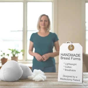 May include: Two white, handmade breast forms and a pile of white cotton on a wooden table. A tag reads "HANDMADE Breast Forms" with the words "Lightweight, Cotton, Breathable" and "Designed by a Mastectomy Patient".
