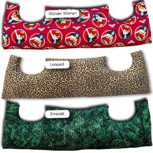 May include: Three neck pillows with different patterns. The top pillow is red with Wonder Woman graphics. The middle pillow has a leopard print. The bottom pillow is emerald green with a textured pattern. Each pillow has a unique shape.