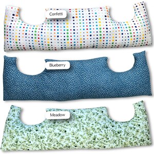 May include: Three long, curved pillows with cut-out sections. The top pillow has a white background with colourful polka dots. The middle pillow is navy blue with white dots. The bottom pillow has a light blue background with green leaf patterns. Each pillow has a label.