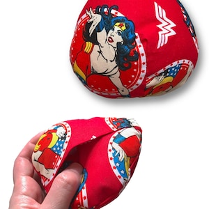 Puede incluir: Bolsa de tela roja con un dise&ntilde;o de c&oacute;mic de Wonder Woman. La bolsa presenta un patr&oacute;n repetido de ilustraciones de Wonder Woman y el logotipo de Wonder Woman. La bolsa se sostiene en una mano.