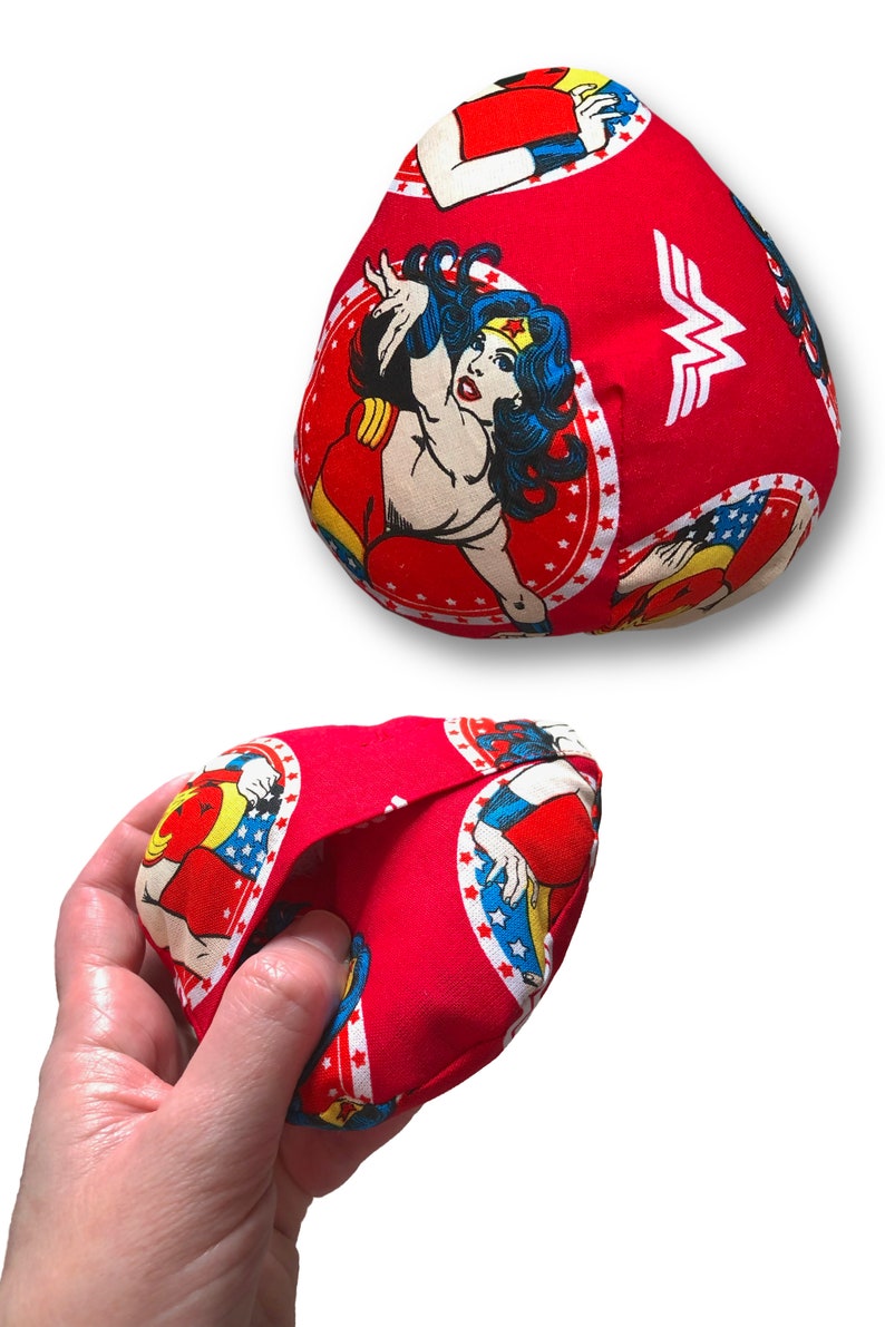 Puede incluir: Bolsa de tela roja con un dise&ntilde;o de c&oacute;mic de Wonder Woman. La bolsa presenta un patr&oacute;n repetido de ilustraciones de Wonder Woman y el logotipo de Wonder Woman. La bolsa se sostiene en una mano.