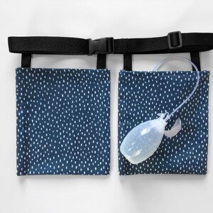 Puede incluir: Un soporte para bolsa de drenaje m&eacute;dico con dos bolsillos rectangulares de tela azul marino con un patr&oacute;n de rayas blancas. Los bolsillos est&aacute;n unidos a un cintur&oacute;n de nailon negro ajustable con hebilla de pl&aacute;stico. Se ve un drenaje de bulbo de pl&aacute;stico transparente.