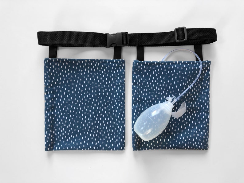 Puede incluir: Un soporte para bolsa de drenaje m&eacute;dico con dos bolsillos rectangulares de tela azul marino con un patr&oacute;n de rayas blancas. Los bolsillos est&aacute;n unidos a un cintur&oacute;n de nailon negro ajustable con hebilla de pl&aacute;stico. Se ve un drenaje de bulbo de pl&aacute;stico transparente.