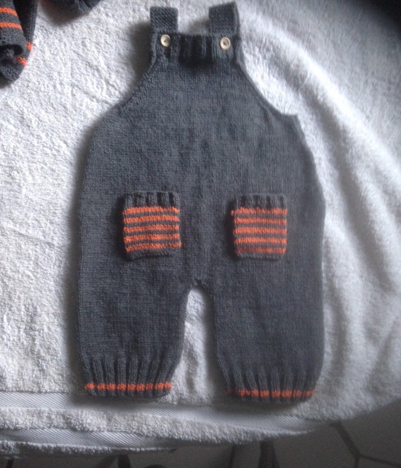 Hand knitted baby dungarees with striped patch pockets detail with over shoulder wooden fastenings also available with matching cardigan.
