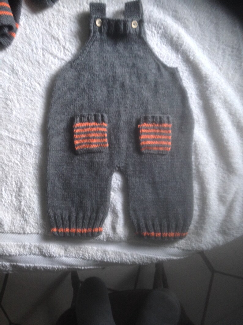 Hand knitted baby dungarees with striped patch pockets detail with over shoulder wooden fastenings also available with matching cardigan.