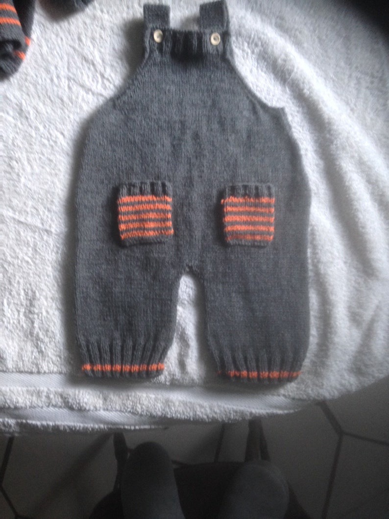 Hand knitted baby dungarees with striped patch pockets detail with over shoulder wooden fastenings also available with matching cardigan.
