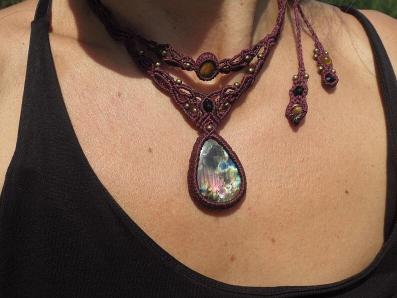 May include: A maroon macrame necklace with a teardrop-shaped iridescent labradorite gemstone pendant. The necklace features smaller tiger's eye and black beads.