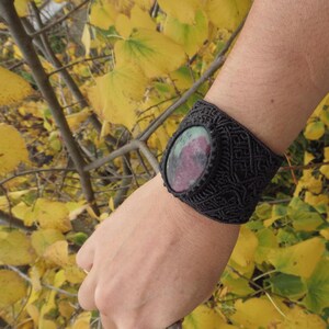 Puede incluir: Un brazalete de macramé negro con una piedra ovalada verde y rosa en el centro.