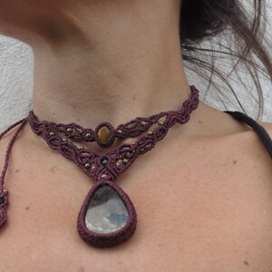 May include: A dark purple macrame choker necklace with a teardrop-shaped pendant featuring a gray and black stone. The necklace has a smaller round stone in the center of the choker.