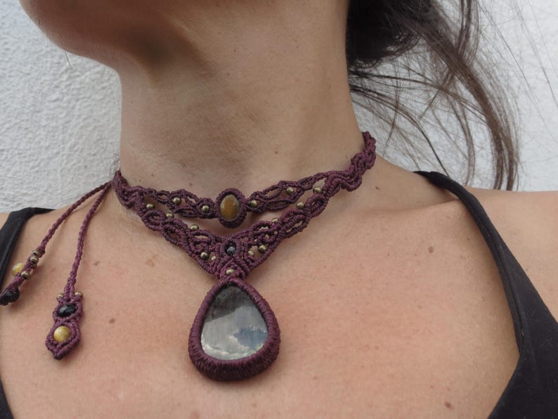 May include: A dark purple macrame choker necklace with a teardrop-shaped pendant featuring a gray and black stone. The necklace has a smaller round stone in the center of the choker.