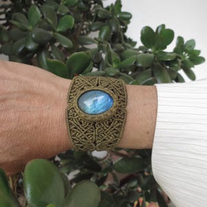 Könnte beinhalten: Handgefertigtes Armband aus olivgrünem Makramee mit einem ovalen Labradorit-Stein. Der Stein zeigt blaue und grüne Schimmer. Das Armband wird am Handgelenk getragen und zeigt die komplizierte Knotenmusterung und die natürliche Schönheit des Edelsteins.