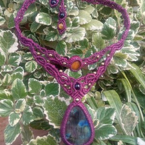 May include: A purple macrame necklace with a teardrop-shaped labradorite gemstone pendant. The necklace is accented with small gold and black beads.