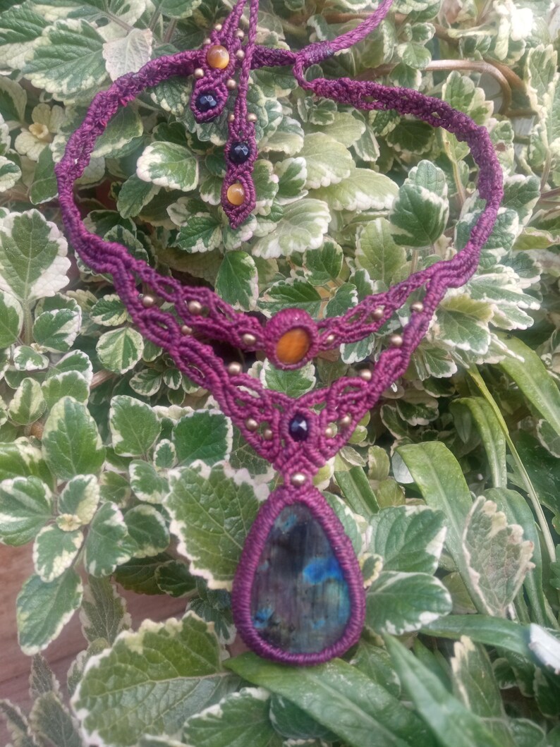 May include: A purple macrame necklace with a teardrop-shaped labradorite gemstone pendant. The necklace is accented with small gold and black beads.