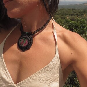 Può includere: Primo piano di una donna che indossa un top bianco con spalline e una collana in macramé nera con un ciondolo di pietra preziosa rosa e verde.