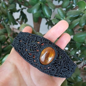 Può includere: Braccialetto in macramè nero con una pietra ovale color ambra. Il braccialetto presenta un intricato design intrecciato. Lo sfondo è una lussureggiante pianta verde.