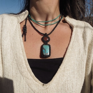 Puede incluir: Un collar con un colgante de piedra turquesa y marrón con un cordón negro. El colgante es una piedra turquesa rectangular con una piedra marrón redonda más pequeña encima. El collar lo lleva una persona con un suéter blanco.