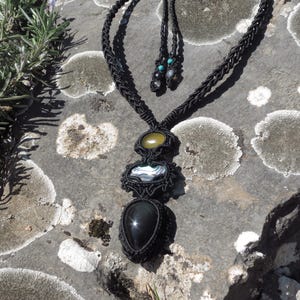 Puede incluir: Un collar de macramé negro con tres colgantes de piedras preciosas. El colgante superior es una piedra ovalada de color amarillo claro, el del medio es una piedra rectangular blanca y el inferior es una piedra negra en forma de lágrima. El collar se muestra sobre una superficie de piedra gris.