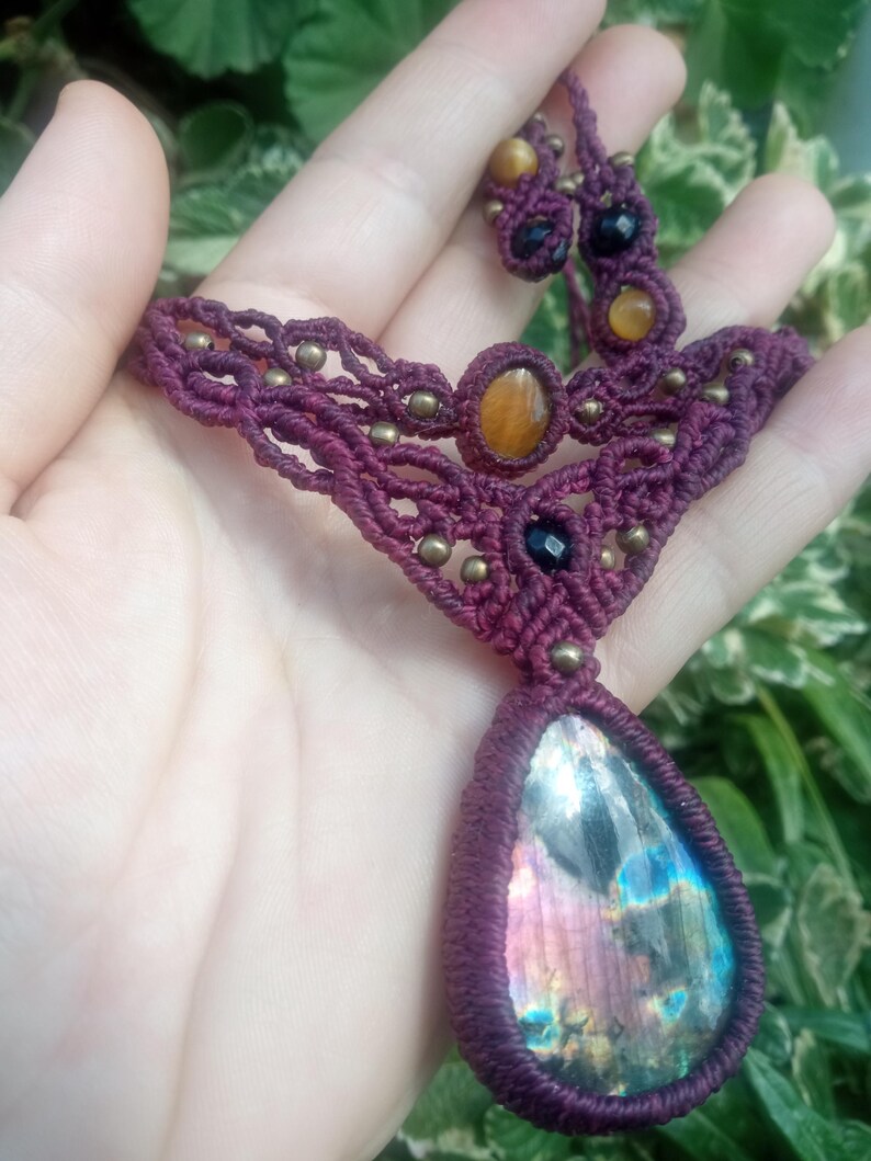 May include: A maroon macrame necklace with a large iridescent labradorite gemstone pendant. The necklace also features smaller tiger's eye and black onyx beads.