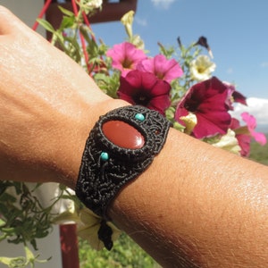 May include: A black macrame bracelet with a red carnelian stone and turquoise accents. The bracelet is worn on a person's wrist.