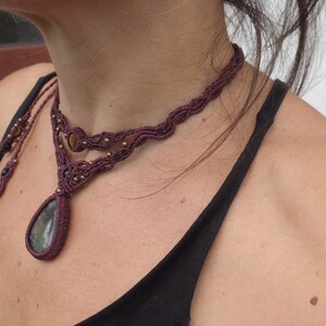 May include: A close-up of a woman wearing a layered macrame necklace with a large, teardrop-shaped gemstone pendant. The necklace is made of dark brown cord and features small beads.