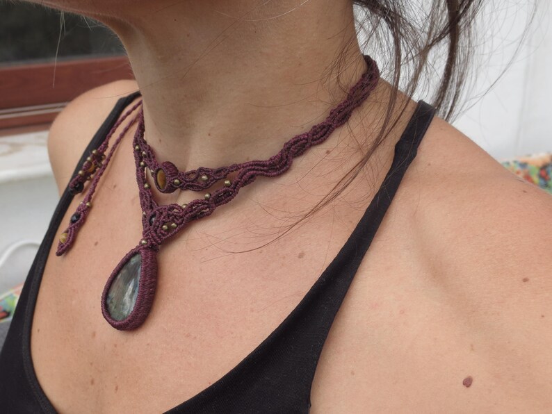 May include: A close-up of a woman wearing a layered macrame necklace with a large, teardrop-shaped gemstone pendant. The necklace is made of dark brown cord and features small beads.