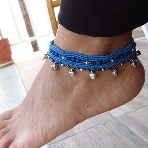 May include: Blue macrame anklet with small silver bells dangling from the bottom. The anklet is adorned with small beads in shades of blue, green, and silver. The anklet is worn on an ankle.