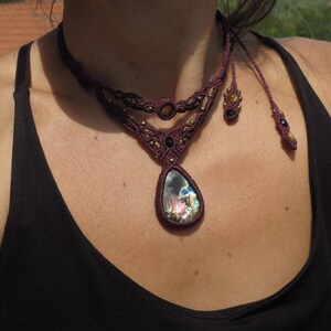May include: A close-up of a woman wearing a dark brown top with a layered macrame necklace featuring a large iridescent gemstone pendant. The necklace has small gold and black beads.