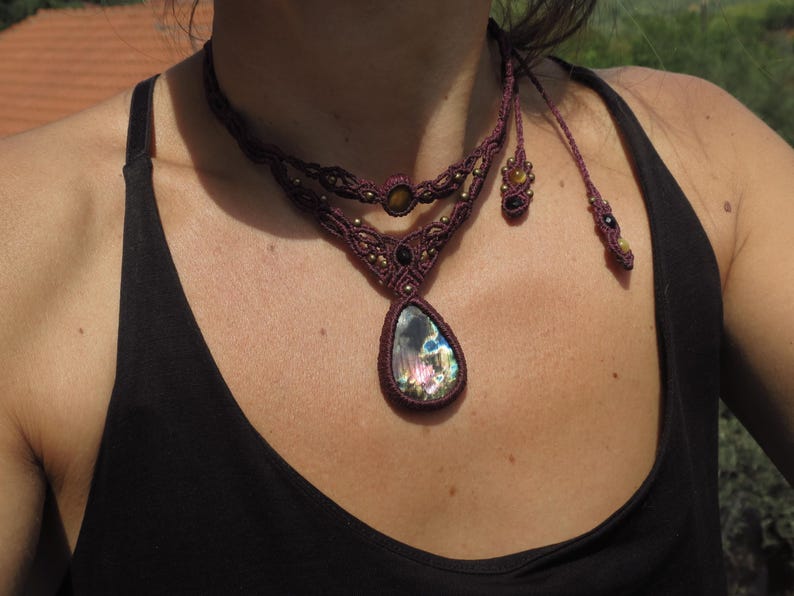 May include: A close-up of a woman wearing a dark brown top with a layered macrame necklace featuring a large iridescent gemstone pendant. The necklace has small gold and black beads.
