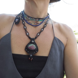 Puede incluir: Conjunto de collares de varias capas con un colgante de macramé negro con una gran piedra ovalada en tonos rosa y verde. El conjunto incluye un colgante a juego y varios collares gargantilla con cuentas en azul, negro y otros colores.