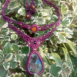 May include: A purple macrame necklace with a teardrop-shaped labradorite gemstone pendant. The necklace features smaller gemstone accents in shades of brown, black, and orange.