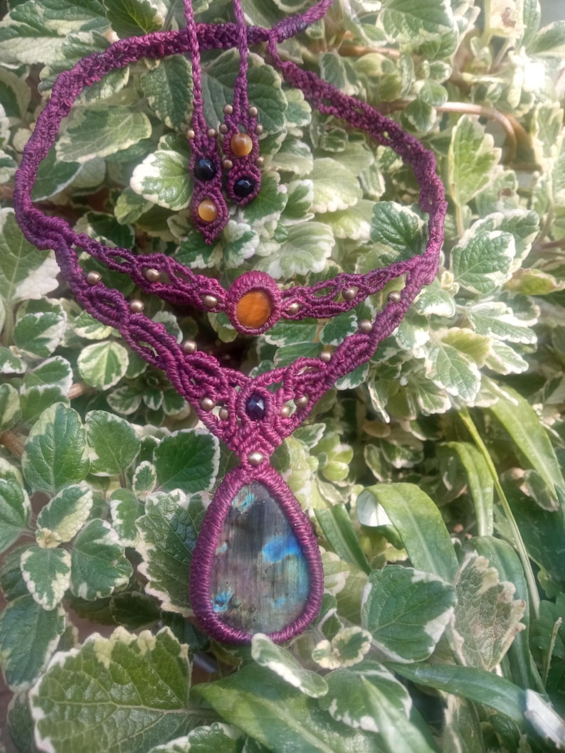 May include: A purple macrame necklace with a teardrop-shaped labradorite gemstone pendant. The necklace features smaller gemstone accents in shades of brown, black, and orange.