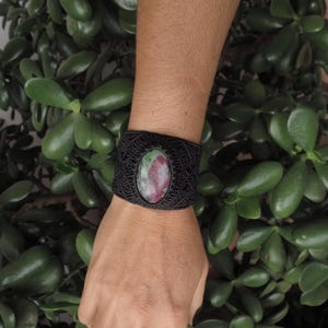 Könnte beinhalten: Ein schwarzes Lederarmband mit einem grün-rosa Edelstein. Das Armband ist mit einem Knotenmuster gewebt.